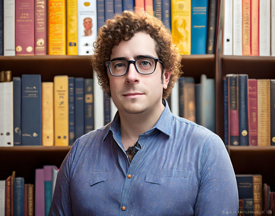 Curly-Haired Person in Blue Shirt with Glasses Standing by Bookshelf