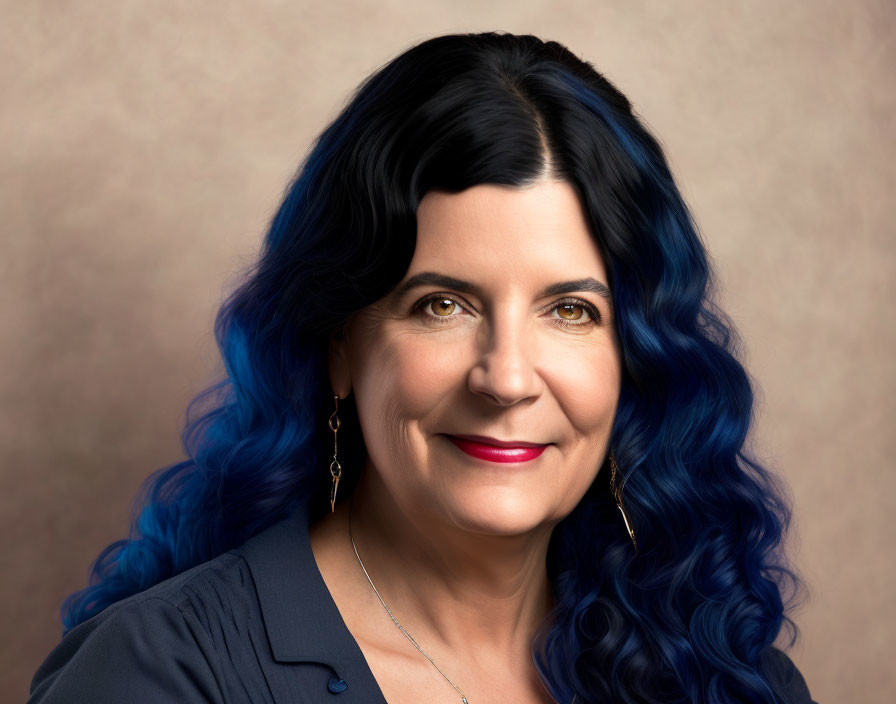 Smiling woman with black and blue hair, earrings, and dark blouse on beige background