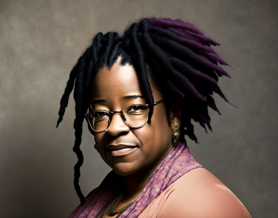 Portrait of person with purple-tinted dreadlocks and glasses in pink garment against brown background