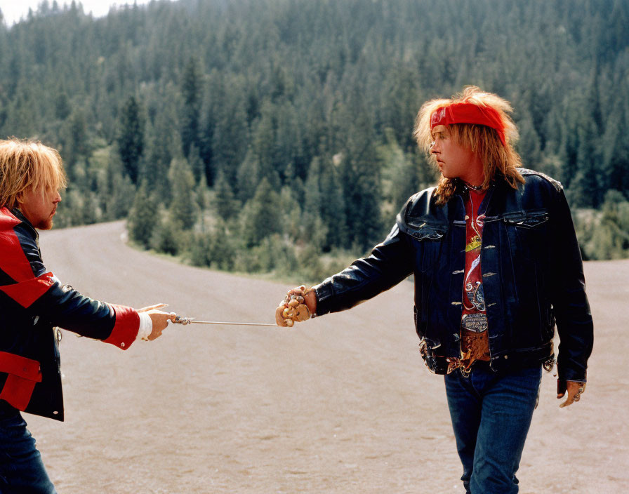 Two people in casual attire playfully sword fight with sticks on forest road