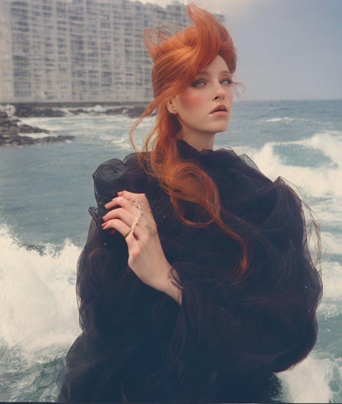 Redhead woman in black outfit by the sea with waves and high-rise building.