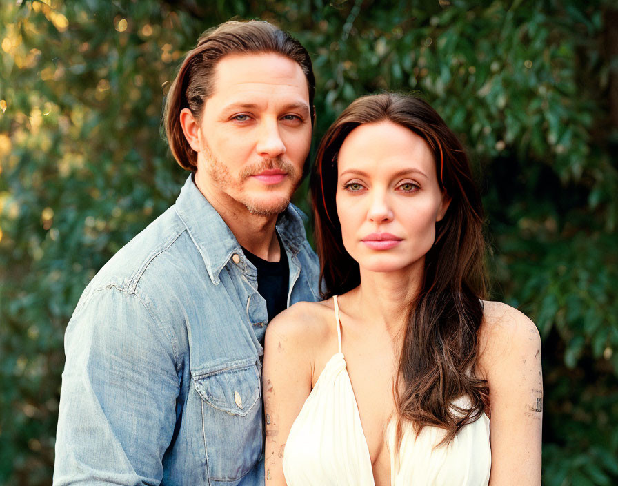 Brown-haired man and woman in denim and white tops pose against green foliage