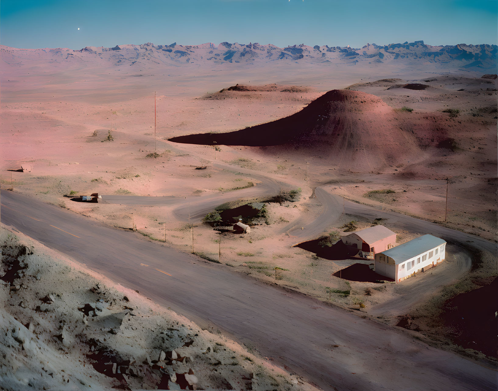 Desert landscape at twilight with winding roads, lit building, mountains, and starry sky