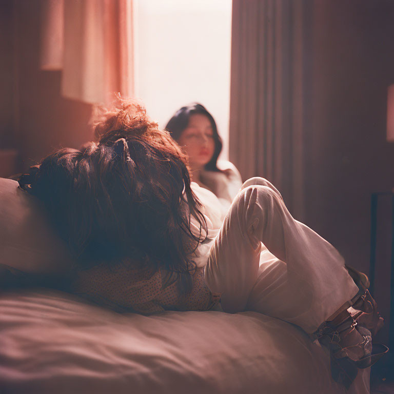 Woman lying on bed with person in background in warm-lit room