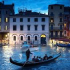 Night scene: Boats on reflective water, illuminated traditional buildings, deep blue sky.