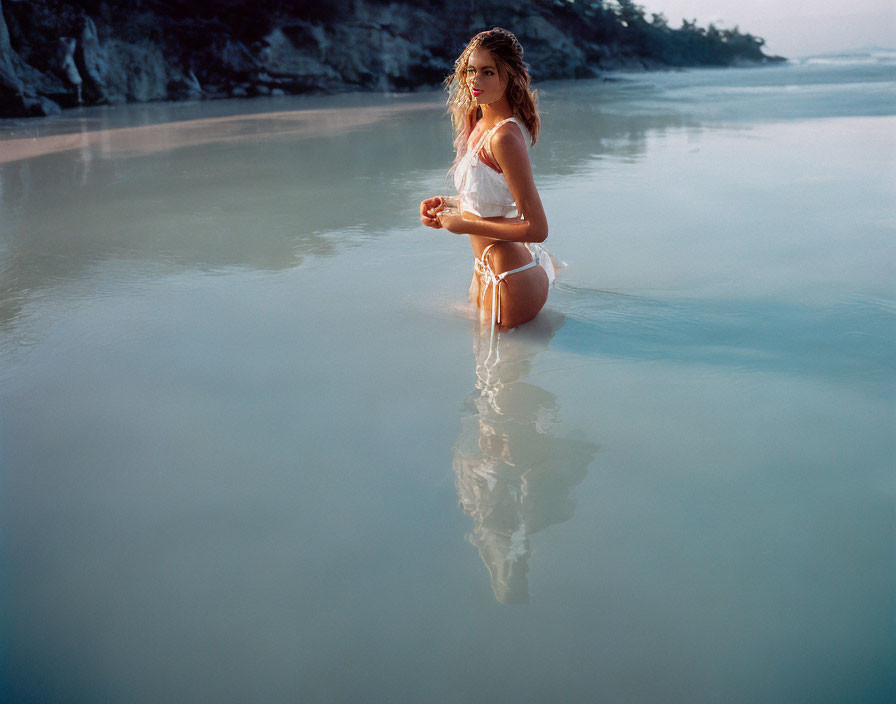 Woman kneeling in shallow ocean water at twilight with reflection visible