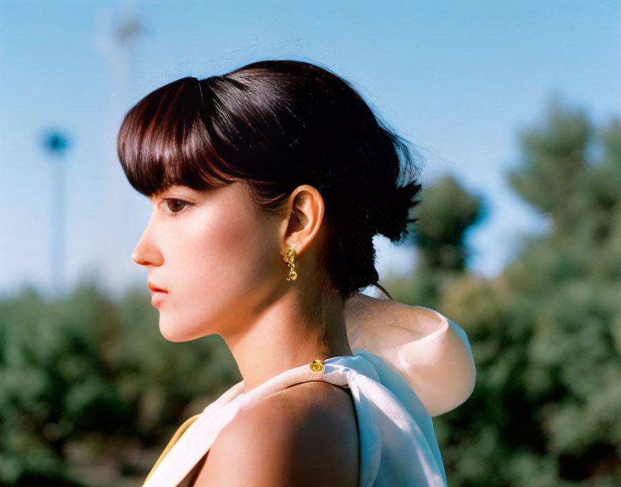 Woman with short hair in yellow dress and earrings, against trees and blue sky.