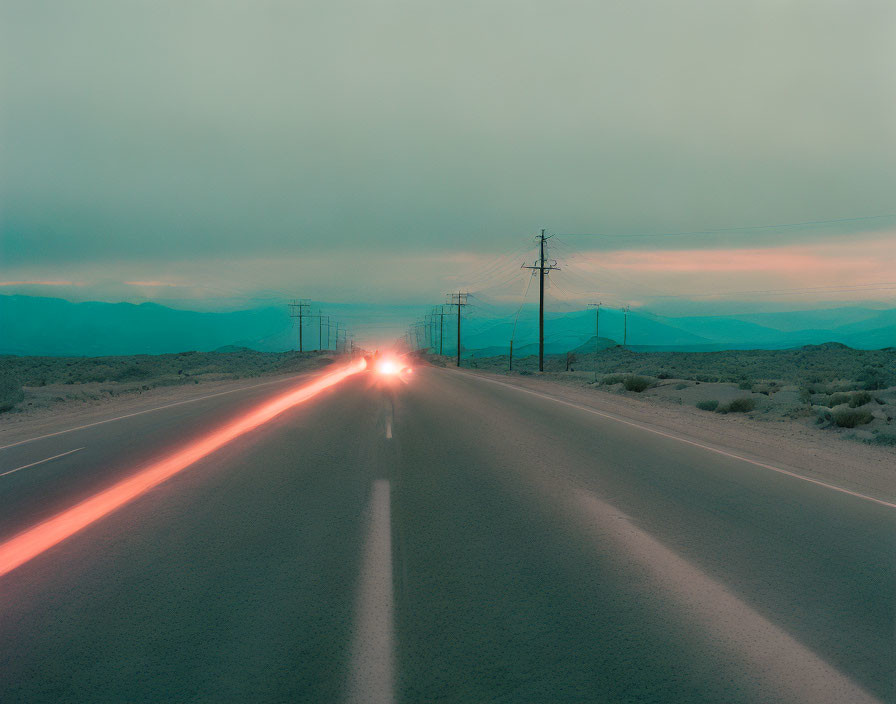 Desolate highway at dusk with car light trails and misty mountains.