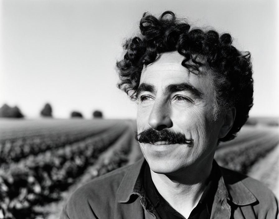 Monochrome portrait of smiling man with curly hair and thick mustache in open field landscape