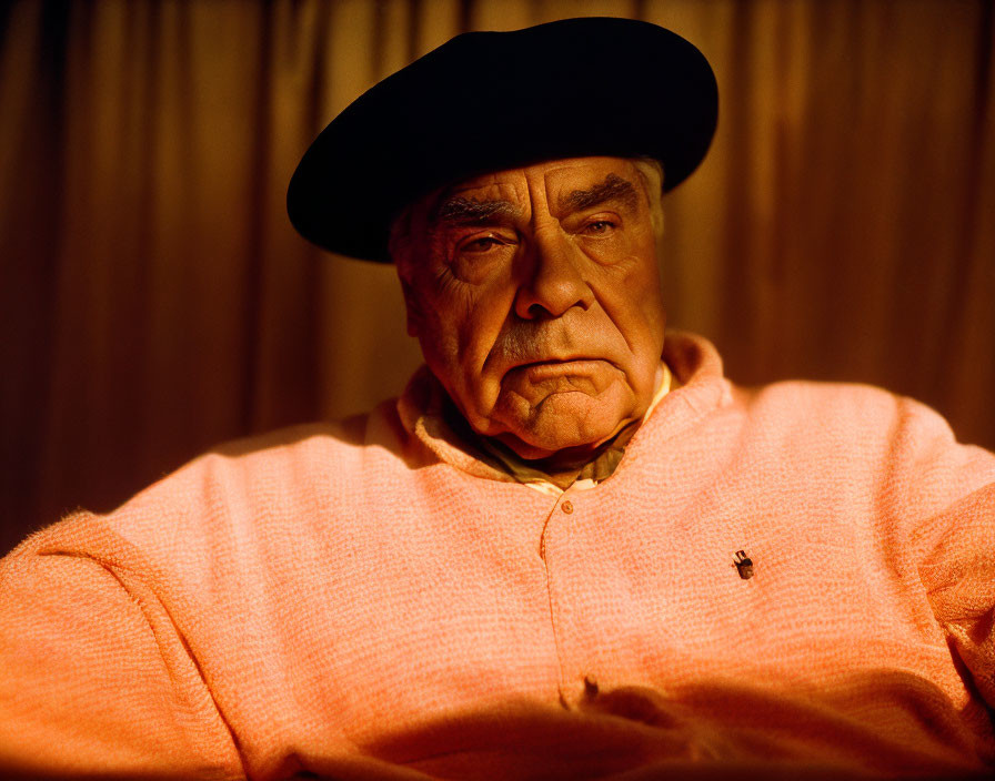 Elderly man in black beret and peach sweater sits thoughtfully in warmly lit room