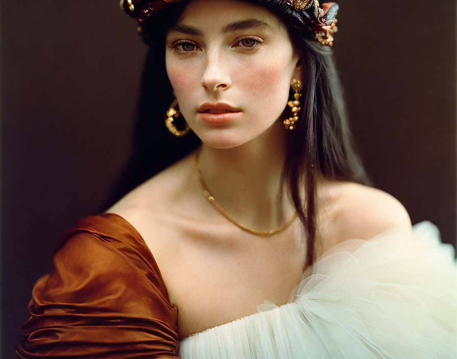 Dark-haired woman in white tulle dress with pearl necklace and gold earrings on brown backdrop