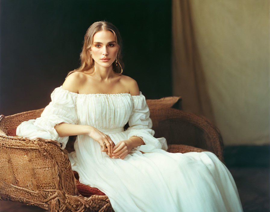 Woman in off-shoulder white dress sitting elegantly on wicker chair against dark backdrop