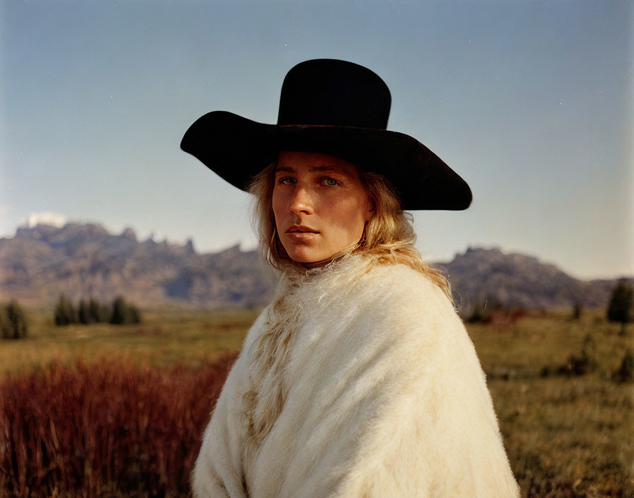 Person with shoulder-length hair in black hat and white fur coat in field with mountains.