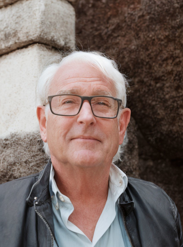 Elderly man with white hair and glasses smiling by stone wall