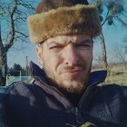 Bearded man with blue eyes in fur hat, rural setting with barns and tractor