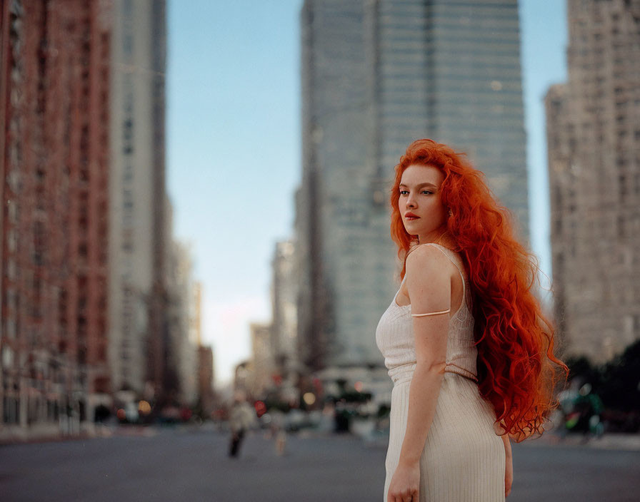 Red-haired woman in white dress on urban street with tall buildings.