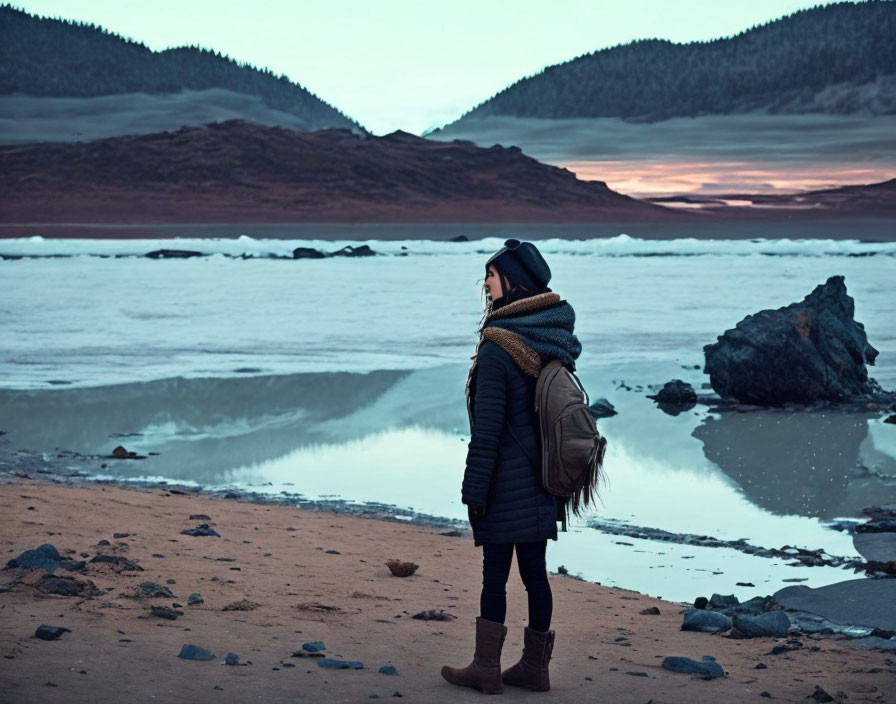 Person in warm clothing by frozen lake at twilight with scenic hills and dusky sky