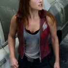 Red-haired woman in grey tank top and red leather vest with holster and gun gazes sideways