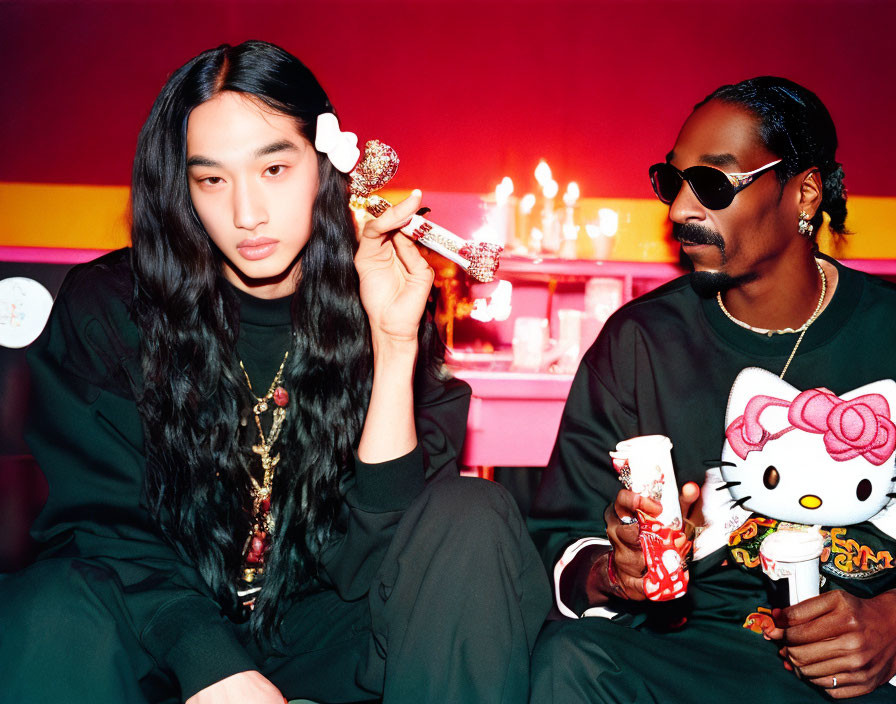 Man and woman posing with Hello Kitty items and candles in stylish attire against red backdrop.