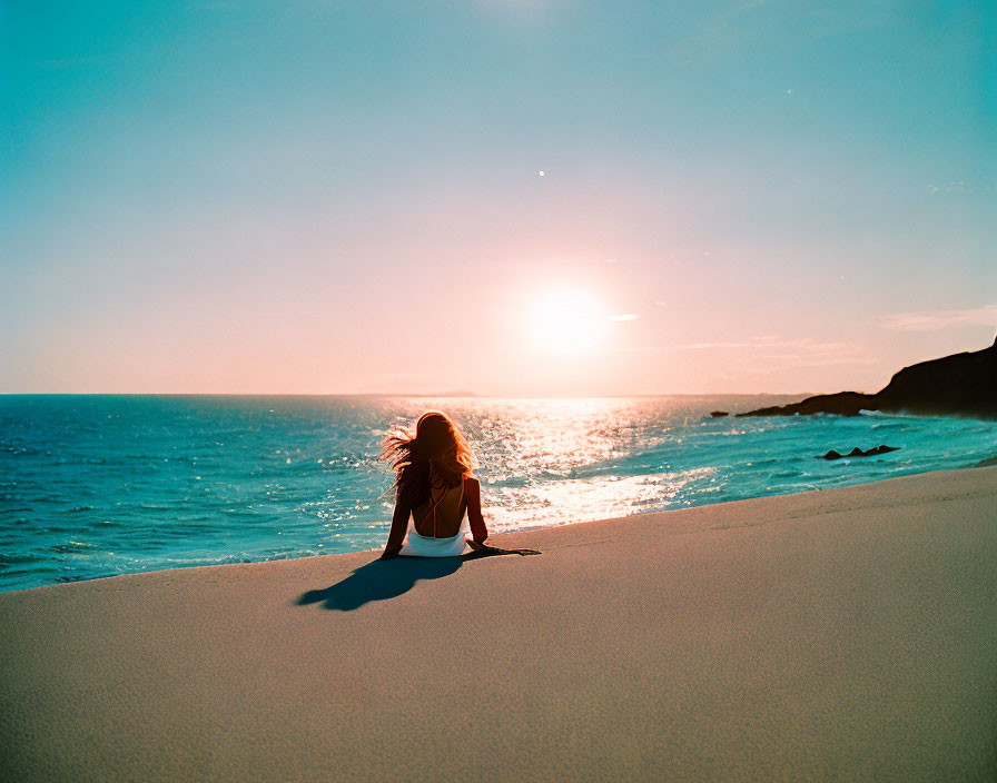 Person watching sunset on sandy beach with ocean waves and clear sky