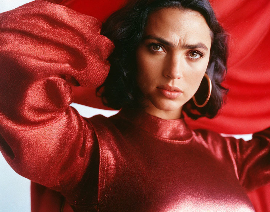 Dark-haired woman in shiny red top with voluminous sleeves and gold hoop earrings posing against red backdrop