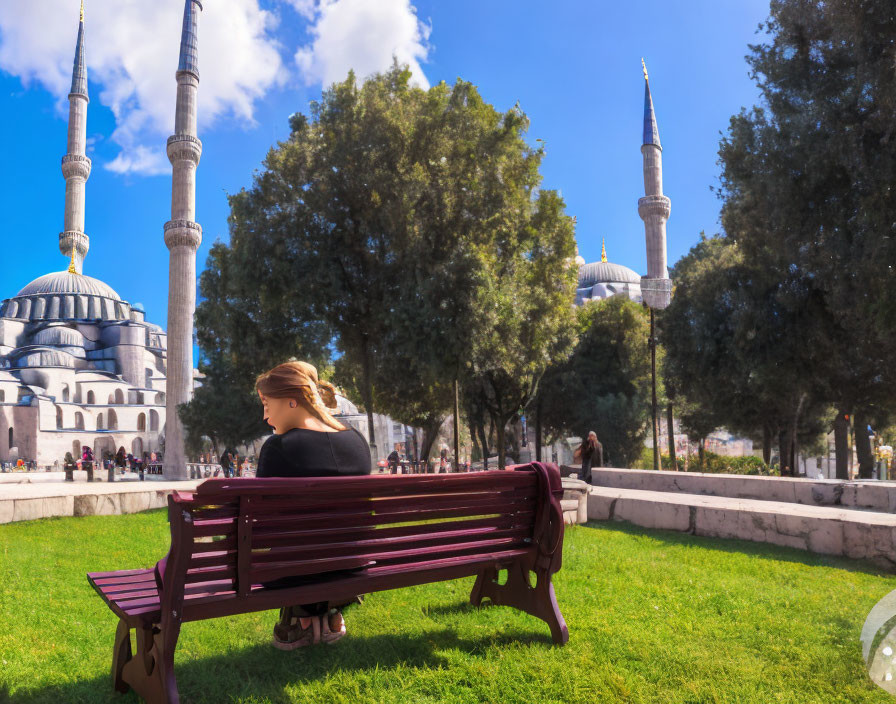 Person sitting on bench in park facing mosque with minarets under blue sky