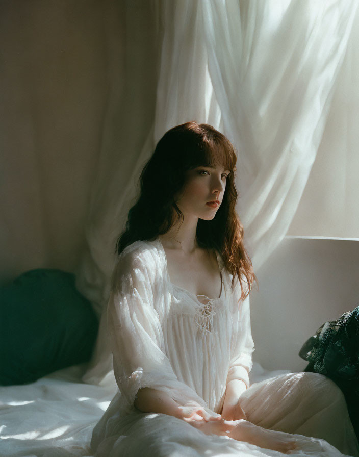 Serene woman in white dress by window with billowing curtains