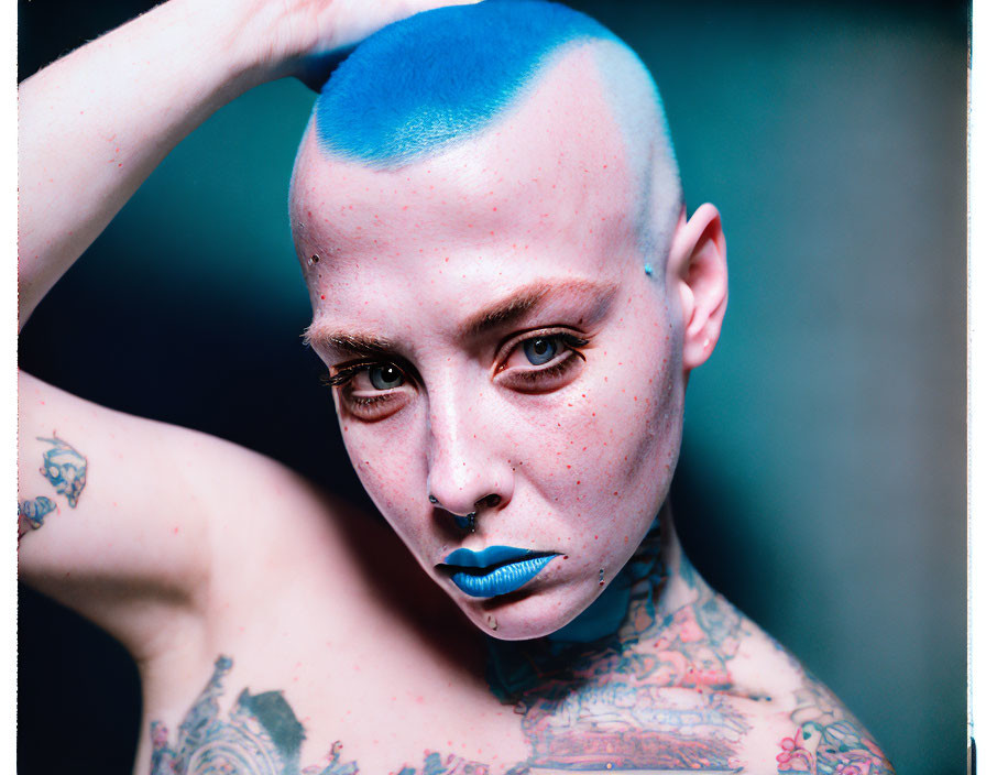 Blue-shaved head person with tattoos and freckles staring at camera