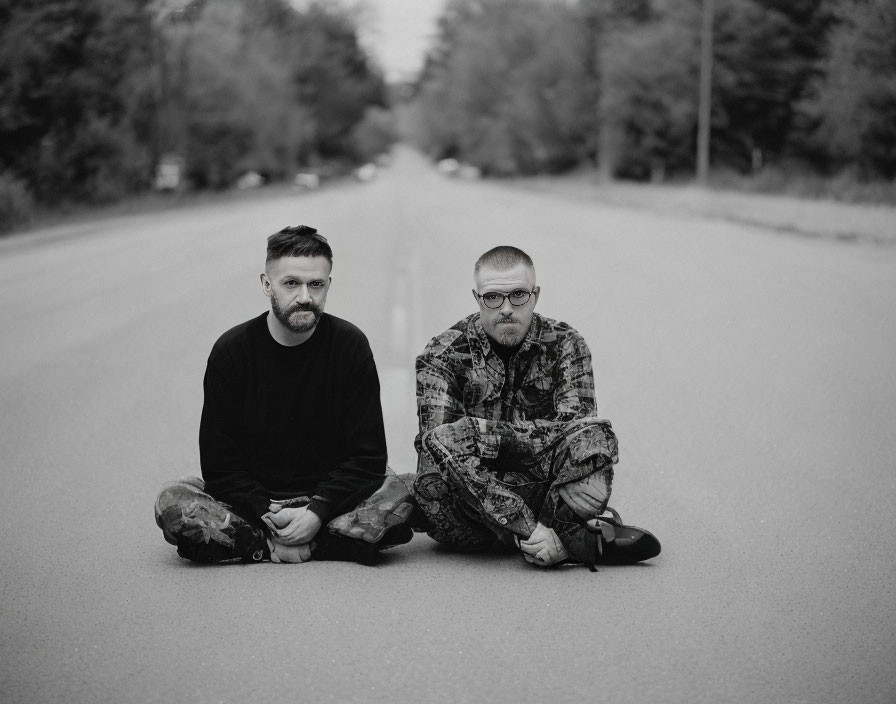 Two men sitting on deserted road, looking solemnly at camera