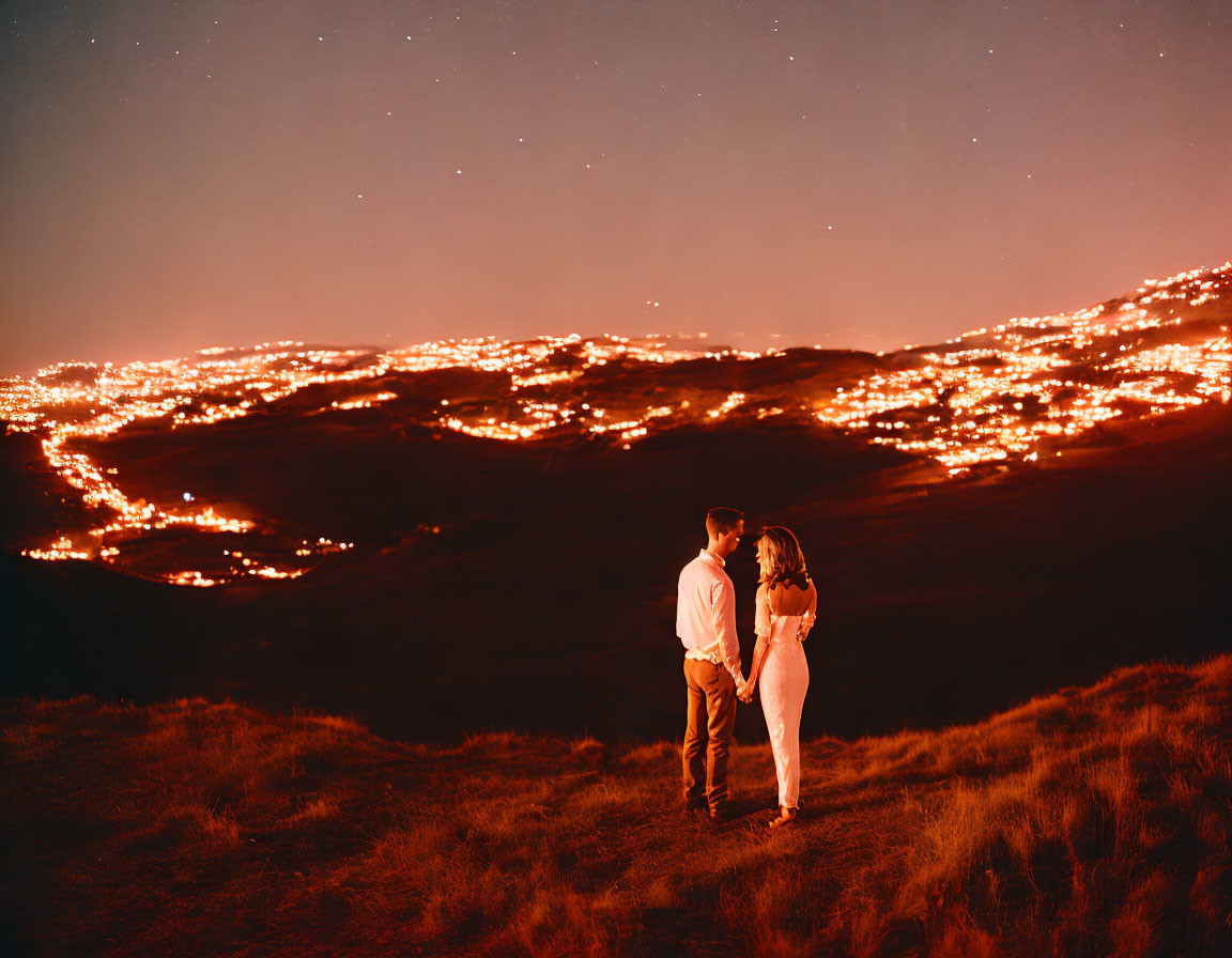 Couple standing on hilltop overlooking cityscape at night