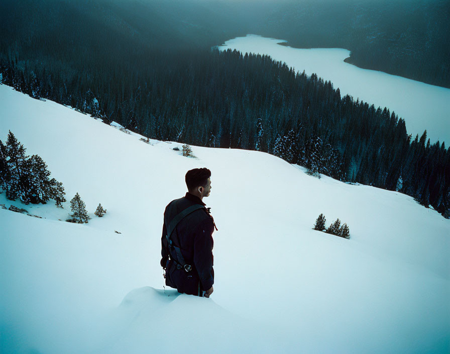 Person with backpack overlooking snowy hill and frozen lake surrounded by forested slopes