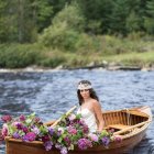 Woman in white dress and hat surrounded by colorful flowers in wooden boat on serene river