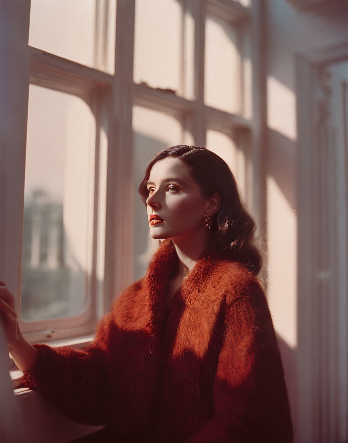 Woman in red fur coat by window in warm sunlight.