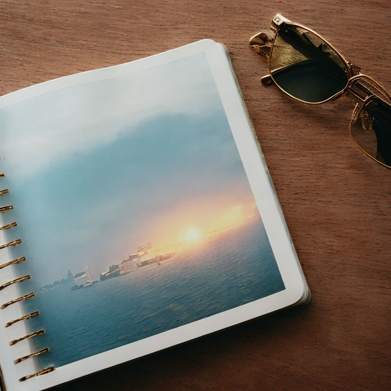 Notebook with sunset seascape photo and sunglasses on wooden surface