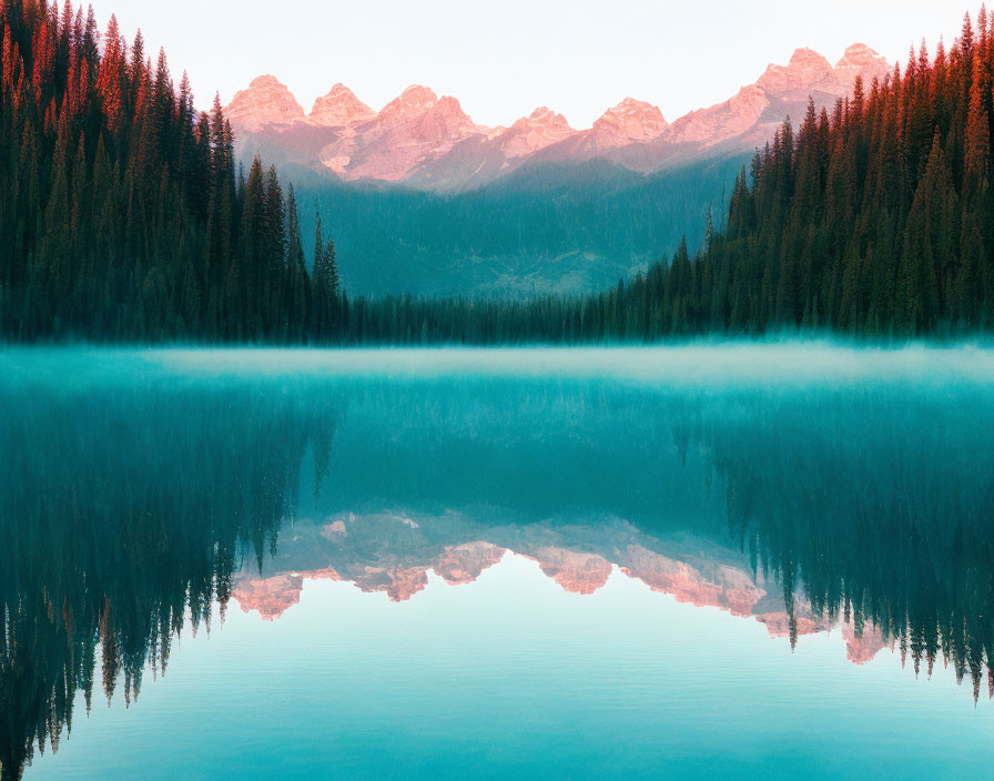 Tranquil lake with misty waters reflecting forest and pink mountain range at dawn.