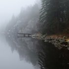 Tranquil lakeside scene with wooden dock, boat, cabin, trees, fog, and mist