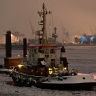 Snowy harbor scene at dusk with moored tugboats and illuminated vessels in calm waters.