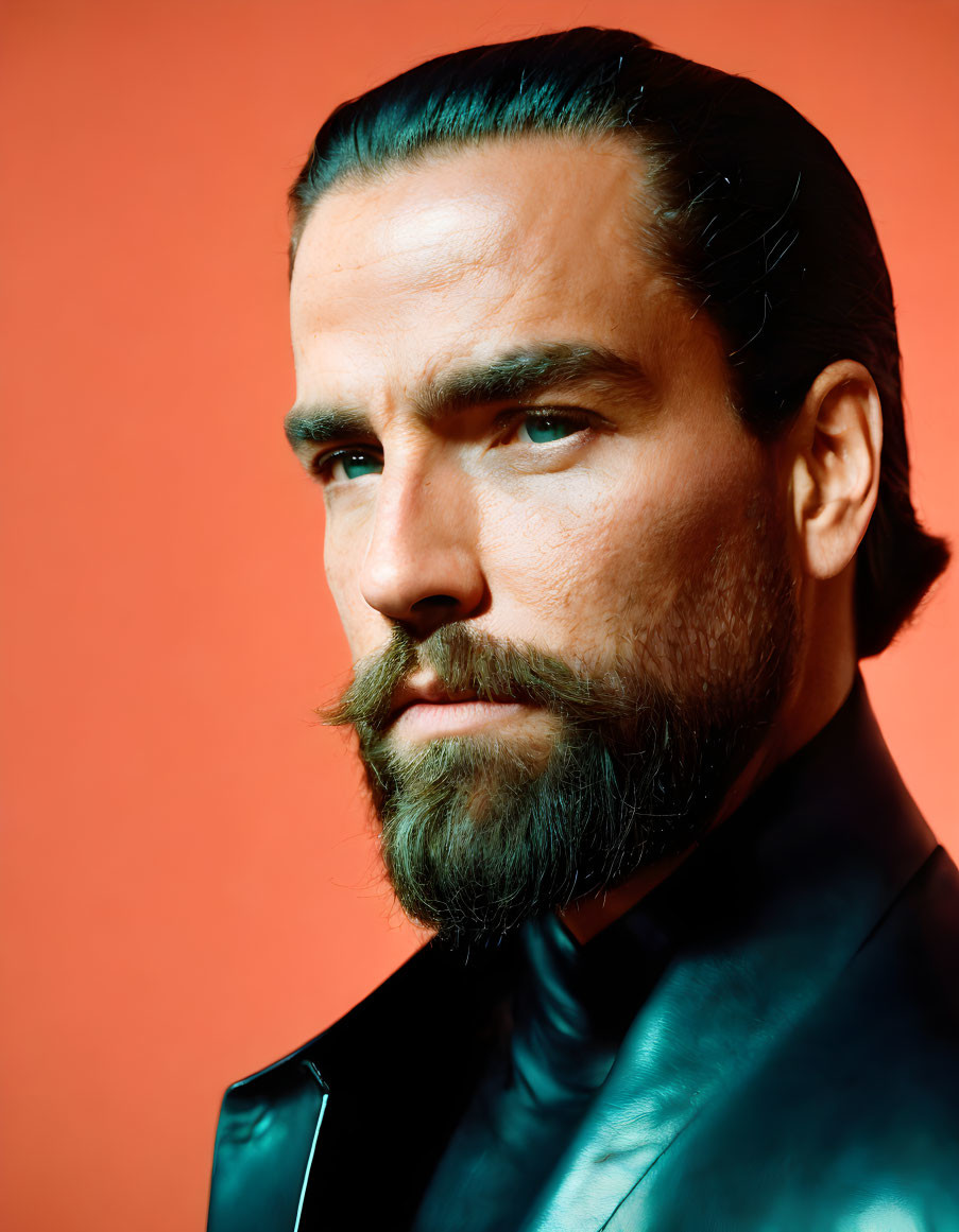 Man with dark slicked-back hair and beard in black leather jacket on orange background