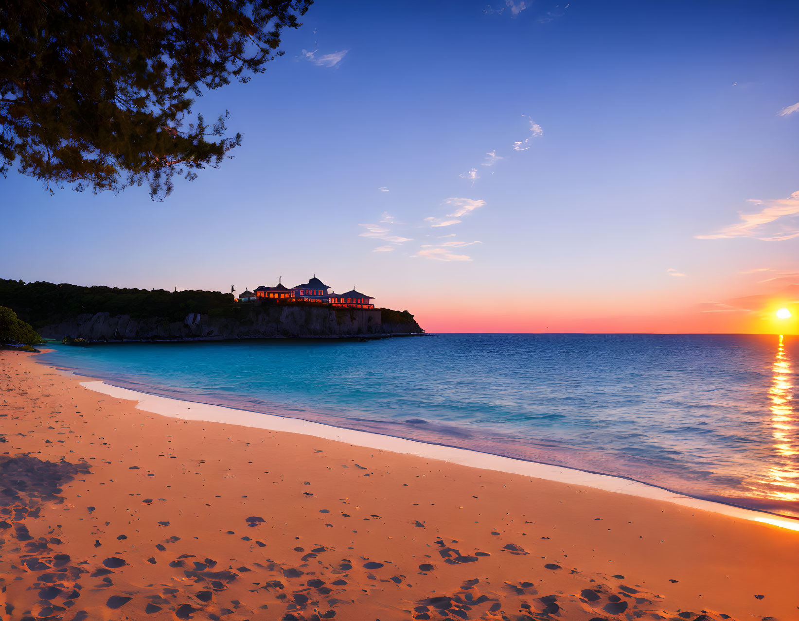 Tranquil beach sunset with clear sky, turquoise water, and grand cliff-top house.