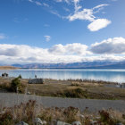 Tranquil landscape with blue lake, snowy mountains, red-roofed cabin, grassy fields