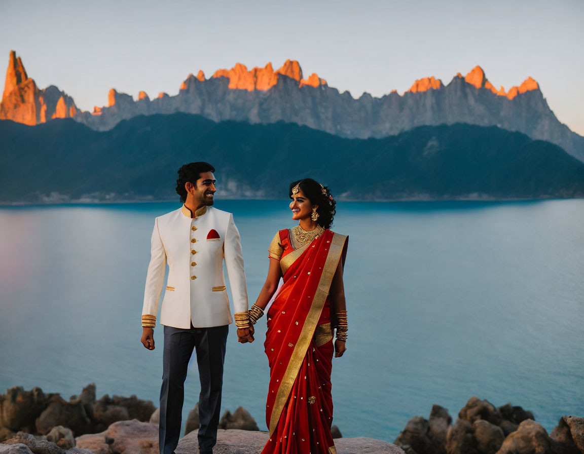 Formal couple holding hands by serene water and mountain peaks at sunset