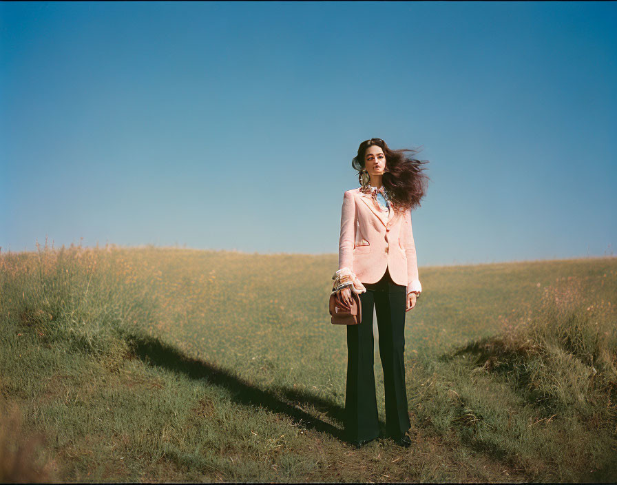 Woman standing in field with hair blowing, wearing pink blazer and black trousers