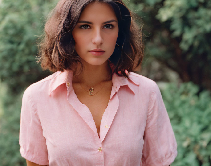 Brown-haired woman in pink shirt and gold necklace against leafy background