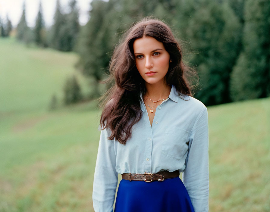 Woman with long brown hair in blue shirt and navy skirt in field with trees and cloudy sky