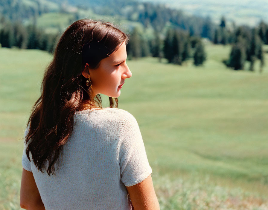 Woman with long brown hair in beige top gazes over green field and hills under blue sky