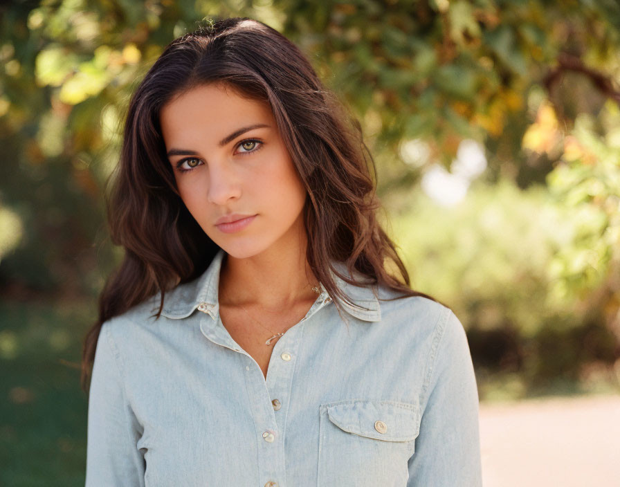 Brown-haired woman in denim shirt standing in front of trees.