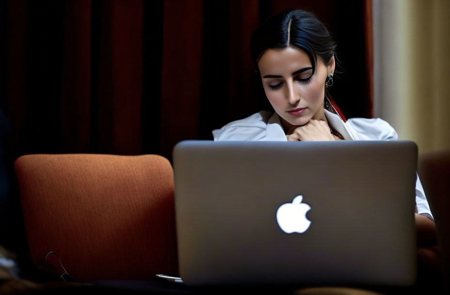 Focused woman working on MacBook in dimly lit room with curtains and orange chair.