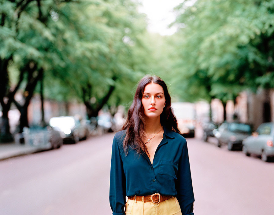 Woman in blue shirt and yellow bottoms on tree-lined street with parked cars.