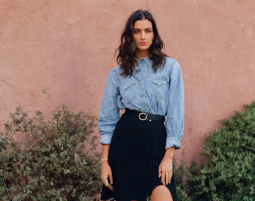 Woman in denim shirt and black skirt against pink wall with green shrubs