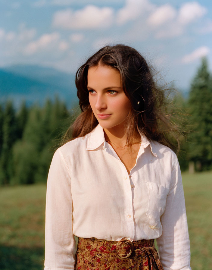 Woman in white blouse and patterned skirt outdoors with trees and mountains.